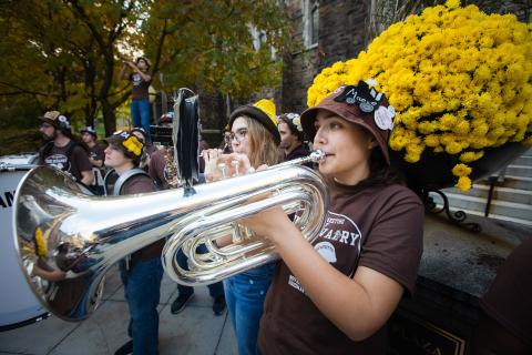 A group of people playing musical instruments outdoors, with one person in the foreground playing a large brass instrument. Bright yellow flowers and autumn trees are visible in the background